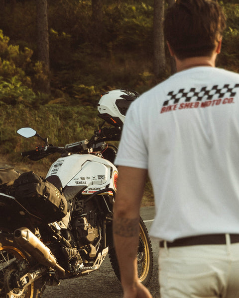 Model wearing Motorcycle Desert Race white t-shirt with a Bike Shed Moto logo, standing next to  a motorcycle in a forest.