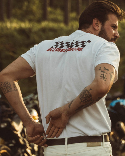 Model wearing Motorcycle Desert Race white t-shirt with a Bike Shed Moto logo, standing in a forest.